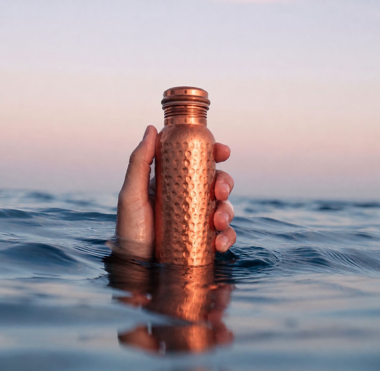 Hand holding a hammered copper Puura bottle emerging from calm sea water at sunset